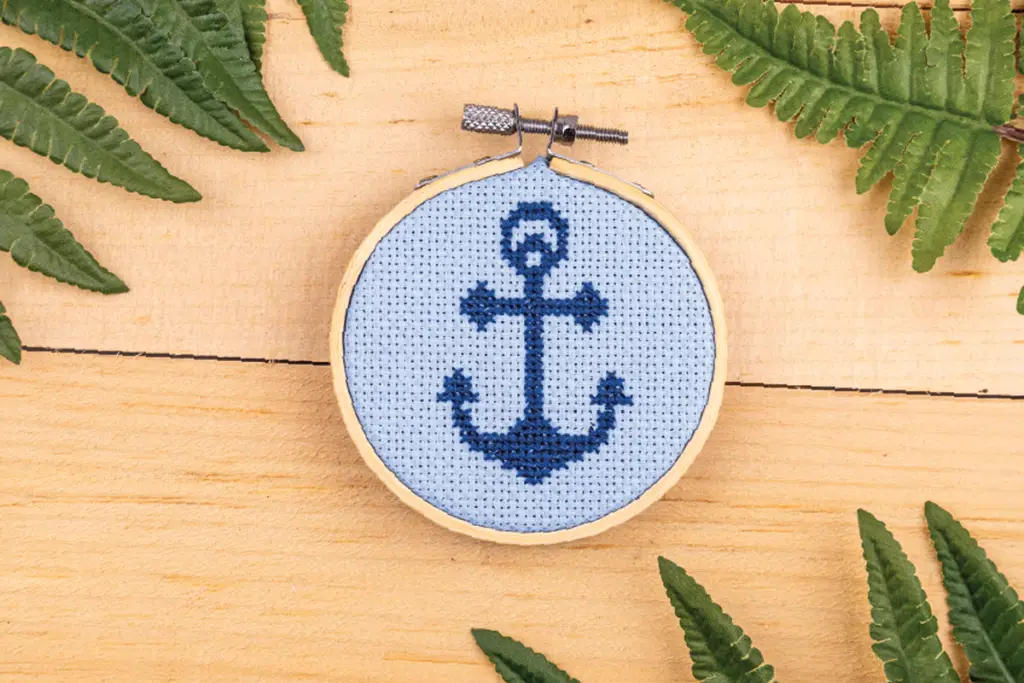Cross Stitched Anchor in Hoop on a light wood background with decorative green leaves