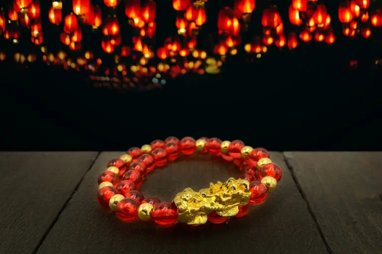 A red and gold beaded bracelet with a large gold dragon charm on a dark wood table with red paper lanterns in the background